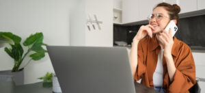 portrait woman working from home with laptop making phone call using smartphone looking 2
